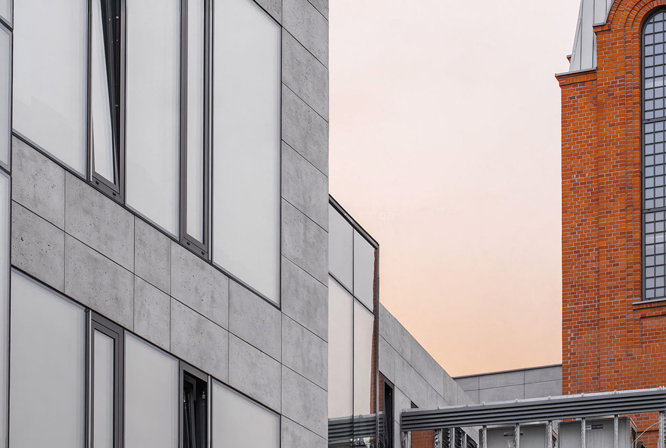Modern building facade featuring sleek concrete panels with a raw, textured finish, combined with large glass windows framed in dark metal. The contemporary design contrasts with the adjacent historic red brick building, creating a visually striking architectural blend.