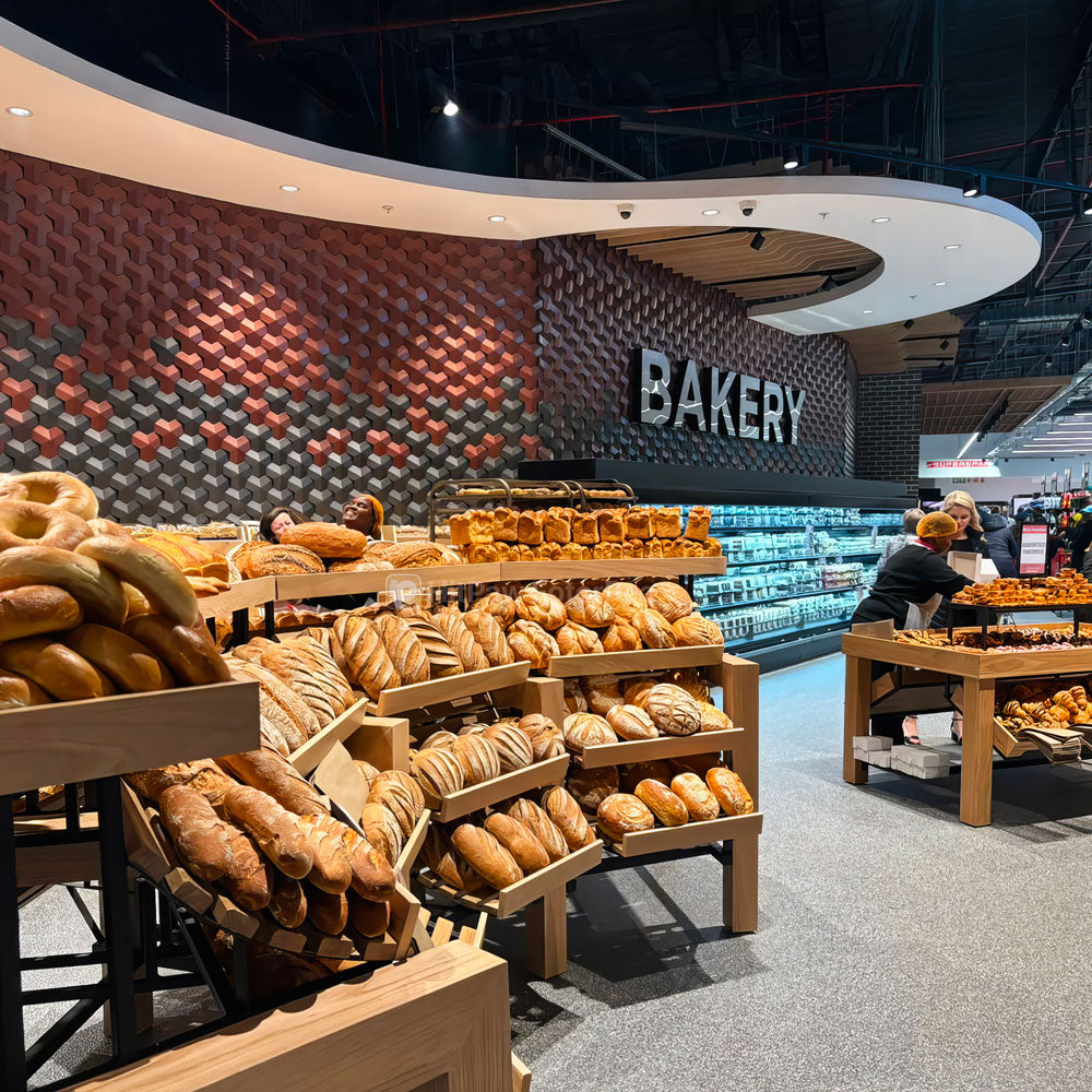 Modern bakery interior showcasing 3D wall tiles with a geometric pattern in red and gray tones, complemented by wooden shelves filled with an assortment of fresh bread and pastries.