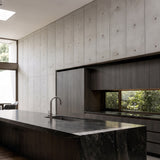 Modern kitchen interior with a dark stone countertop and island, matte gray concrete panel feature wall, dark wood cabinetry, and a curved gooseneck faucet. Windows reveal greenery outside.