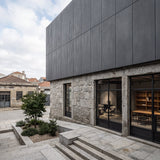 Modern building concrete panel facade with dark gray metal cladding on the upper level and rough stone masonry on the lower level, featuring large black-framed glass windows and a paved courtyard with a small tree and stone steps. - FLOAT concrete