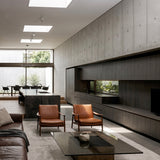 A modern minimalist living room with two mid-century style brown leather armchairs facing a black glass coffee table. The room features a large concrete panel wall with built-in dark wood cabinetry, a black marble kitchen island, and a large window looking out onto a garden.