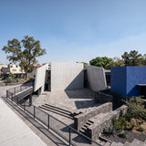 Modernist-style concrete panel wall building with angular walls, a paved courtyard, stone steps, black metal railings, and a vivid blue side structure under a clear blue sky. - FLOAT concrete