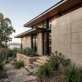 Modern house with stone exterior and glass windows, surrounded by greenery.
