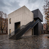 Modernist concrete panels and black bricked building with an external staircase, set in a wet urban courtyard with fallen leaves under an overcast sky. - FLOAT concrete