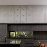 Modern minimalist kitchen interior featuring concrete wall panels, dark wood lower cabinetry, a black stone countertop, and a small window looking out to greenery.