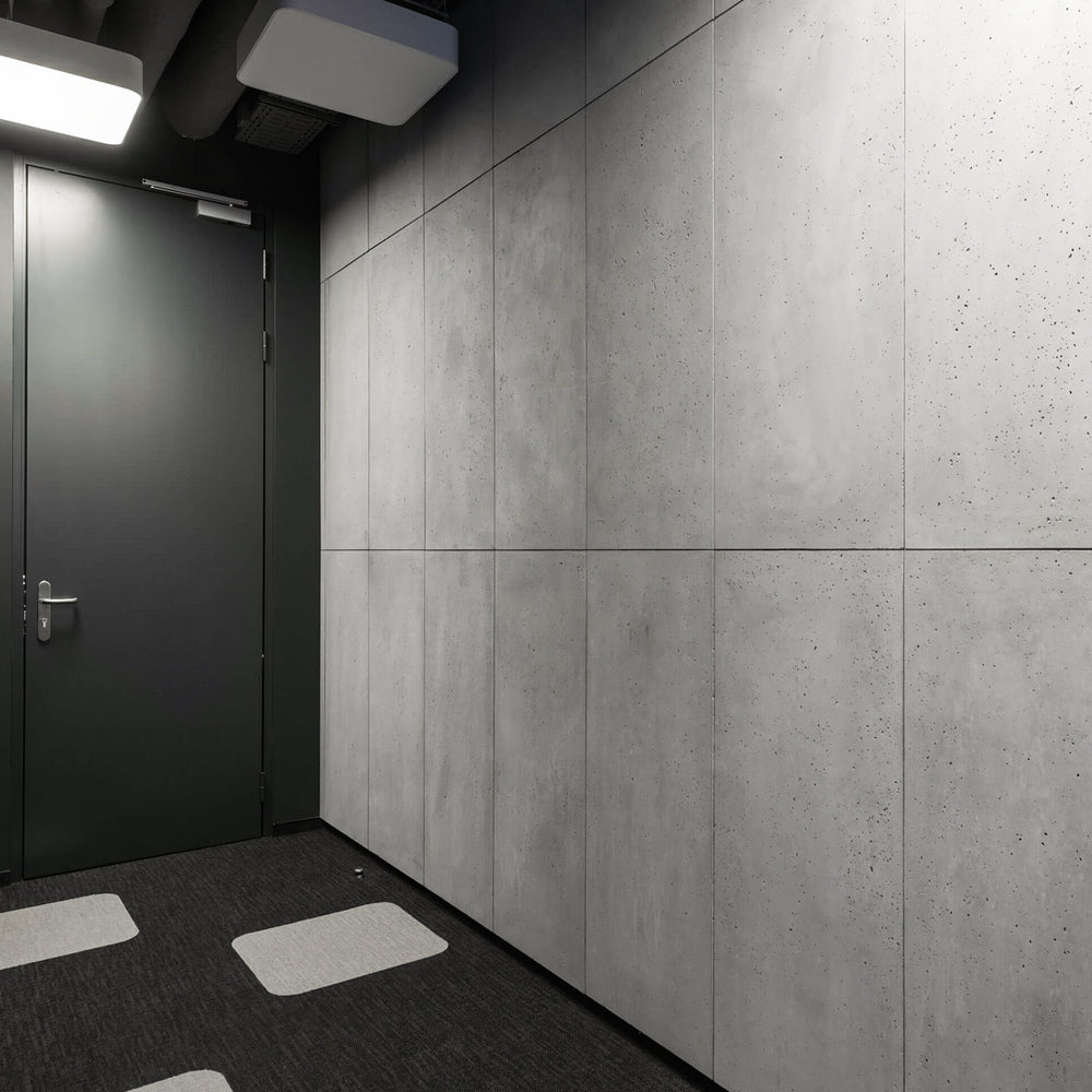 Commercial interior with lightweight grey concrete wall panels and black door, featuring acoustic ceiling elements and dark carpet flooring with light square inlays.